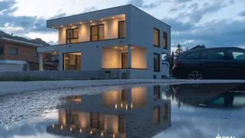 Modernes Einfamilienhaus mit beleuchteter Fassade und Reflexion in einer Pfütze bei Dämmerung.