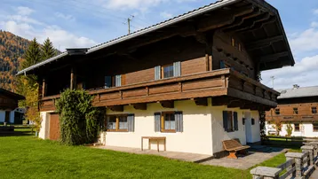 Traditionelles Holzhaus im alpinen Stil mit Balkon, weißer Fassade und grünem Garten vor Bergkulisse.
