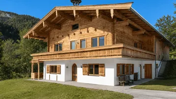 Traditionelles Holzhaus im alpenländischen Stil mit weißem Sockel und Holzbalkonen, umgeben von grüner Natur.