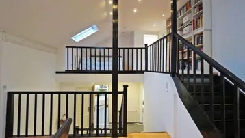 Heller Wohnbereich mit Holztreppe, Galerie und integriertem Bücherregal, durch ein Dachfenster belichtet.