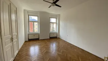 Helles Zimmer mit Fischgrätparkett, hohen Decken und Deckenventilator.