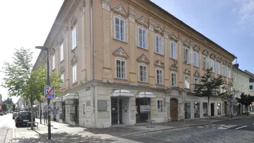 Historisches Gebäude mit aufwendiger Fassade und Geschäften im Erdgeschoss an einer belebten Straße.