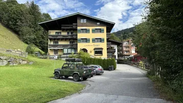 Traditionelles Landhaus in den Bergen mit gelber Fassade, grünen Fensterläden und Parkplätzen.