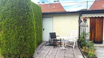 Gemütliche gepflasterte Terrasse mit Sitzgelegenheiten, umgeben von einer hohen Hecke und Blumenbeeten.