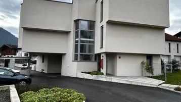 Modernes Gebäude mit klaren Linien und großen Fenstern, umgeben von einer Zufahrt.