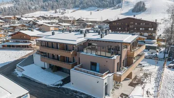 Modernes Gebäude mit Holzbalkonen und schneebedeckten Dächern in alpiner Winterlandschaft.