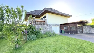 Hausfassade mit Vorgarten, Garageneinfahrt und Carport unter blauem Himmel.