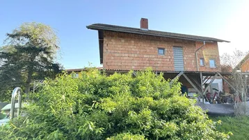 Haus mit Ziegelfassade und Holzbalkenkonstruktion, umgeben von grüner Vegetation unter blauem Himmel.