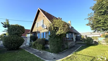 Hausfassade mit Holzverkleidung, Satteldach und gepflegtem Vorgarten unter blauem Himmel.