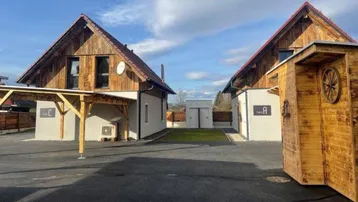 Zwei moderne Häuser mit Holzfassaden, roten Dächern und Carports auf einem gepflasterten Vorplatz.