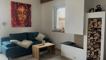 Gemütliches Wohnzimmer mit blauem Sofa, Holztisch und Kamin mit integriertem Holzregal unter Holzbalken.