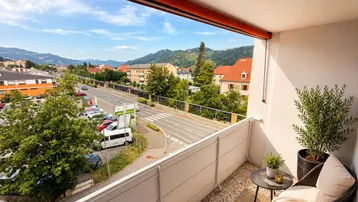 Möblierter Balkon mit Blick auf die Straße, umliegende Gebäude und grüne Hügel.