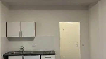 Kleine, funktionale Küchenzeile mit Spüle, Kochfeld und Kühlschrank in einem hellen Raum mit dunklen Fliesen.
