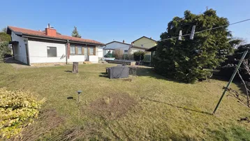 Großer Garten mit Rasenfläche, Bäumen und einem Einfamilienhaus im Hintergrund unter blauem Himmel.