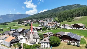 Luftaufnahme eines malerischen Dorfes in den Bergen mit grünen Wiesen und Wäldern unter blauem Himmel.