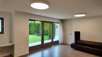 Heller Wohnbereich mit großen Glastüren, die zu einem grünen Garten führen, und einem dunklen Sofa.