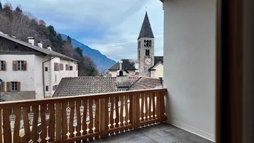Blick vom Balkon auf ein traditionelles Dorf mit Kirchturm und umliegende Berge.