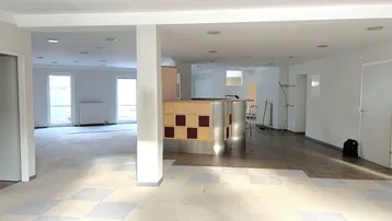 Großer, offener Raum mit einer Bar, kariertem Bodenbelag und mehreren Fenstern, der renovierungsbedürftig ist.