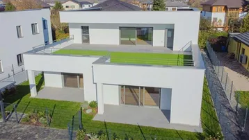 Moderne weiße Architektur mit Flachdach und begrünter Dachterrasse, umgeben von Wohngebäuden.