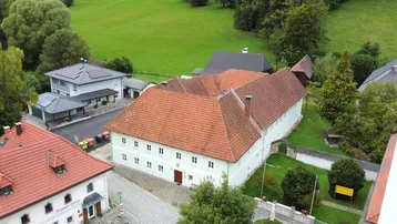 Historisches Gebäude mit rotem Ziegeldach, umgeben von grüner Landschaft und weiteren Häusern.