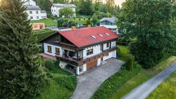 Luftaufnahme eines Einfamilienhauses mit rotem Dach, umgeben von viel Grün und weiteren Häusern.