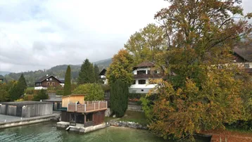Häuser am Seeufer mit herbstlicher Vegetation und Bootsanlegestellen unter bewölktem Himmel.