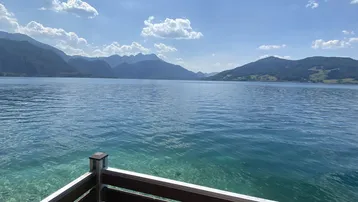 Weitläufiger Blick auf den See und die umliegenden Berge von einem Balkon aus unter blauem Himmel.