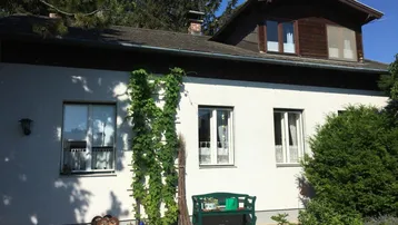 Seitenansicht des Hauses mit weißer Fassade, Kletterpflanzen und einer Gartenbank im Grünen.
