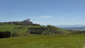 Weitläufige grüne Hügellandschaft mit einem Dorf und einer Kirche auf einem Hügel unter blauem Himmel.