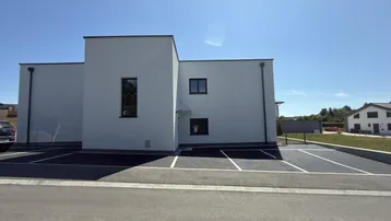 Modernes Neubaugebäude mit weißer Fassade und Flachdach, umgeben von Parkplätzen unter blauem Himmel.