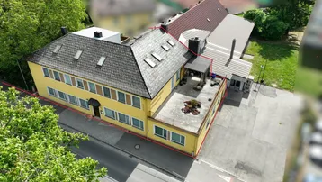 Luftaufnahme eines gelben Gebäudes mit großer Dachterrasse, Schrägdach und umliegendem Grün.