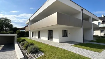 Modernes Mehrfamilienhaus mit weißer Fassade, Balkonen, gepflegtem Vorgarten und Carport unter blauem Himmel.