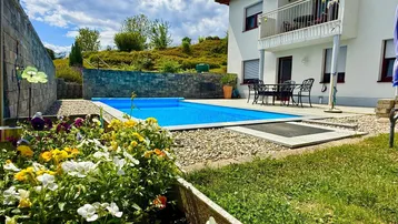 Außenbereich mit Swimmingpool, Terrasse und Balkon, umgeben von blühenden Blumenbeeten und grüner Landschaft.