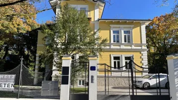 Elegante gelbe Villa mit weißer Fassadenverzierung, umgeben von Bäumen und einem schmiedeeisernen Eingangstor.