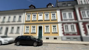 Gelbe Fassade eines Mehrfamilienhauses mit grüner Eingangstür und parkendem Auto auf der Straße.