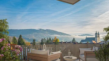 Gerenderter Balkon mit Holzboden, bequemen Möbeln und weitem Blick auf die Berge und eine Kirche.