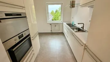 Moderne Küche mit weißen Schränken, integriertem Backofen und Fenster mit Grünblick.