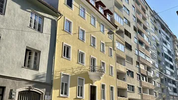 Gelbes Mehrfamilienhaus an einer sonnigen Straße mit parkenden Autos und angrenzenden Gebäuden.