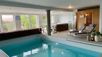 Großzügiger Wellnessbereich mit Innenpool, Whirlpool und Sauna, umgeben von großen Fenstern mit grüner Aussicht.