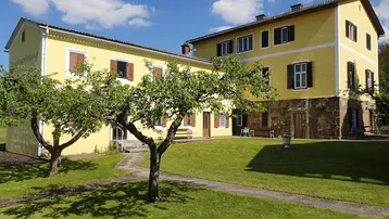 Gut erhaltenes traditionelles Gebäude mit gelber Fassade, umgeben von einem gepflegten Rasen und Bäumen unter blauem Himmel.