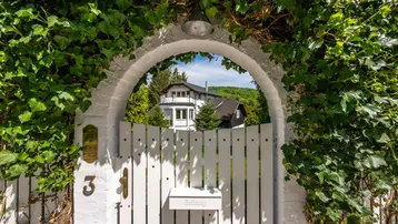 Eingangstor mit üppigem Efeu bewachsen, das den Blick auf die dahinterliegende Villa freigibt.