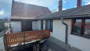 Blick auf die obere Terrasse mit Holzgeländer und angrenzendem Gebäude unter blauem Himmel.
