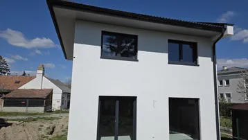 Neubau-Einfamilienhaus mit weißer Fassade und dunklen Fenstern unter blauem Himmel.