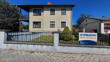 Vorderansicht eines gepflegten Hauses mit Garten, Zaun und einem Verkaufsplakat.