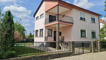 Rosa gestrichenes Einfamilienhaus mit Balkon und Vorgarten, umzäunt von einem Stein- und Metallzaun.