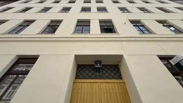 Fassade eines Mehrfamilienhauses mit gelber Eingangstür und vielen Fenstern unter blauem Himmel.