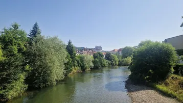 Weitläufiger Flussblick mit grünen Ufern und einer Stadtkulisse in der Ferne unter blauem Himmel.