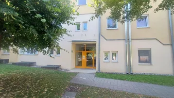 Eingangsbereich eines Mehrfamilienhauses mit heller Fassade, umgeben von Bäumen und Grünflächen.