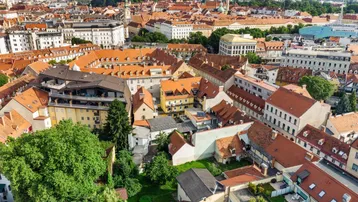 Luftaufnahme der Stadt mit roten Dächern und grünen Hügeln im Hintergrund unter blauem Himmel.