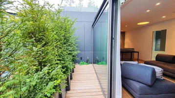 Moderne Holzterrasse mit üppiger Bepflanzung und direktem Zugang zum hellen Innenbereich.